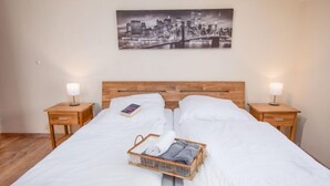 A neatly made bed with white linens, two bedside tables with lamps, a cityscape painting on the wall, and a wicker basket with towels. - Sandburg by Interhome (Norddeich)