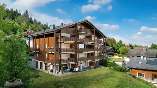 Ein mehrstöckiges Holzgebäude mit Balkonen, umgeben von Grünflächen und blauem Himmel.