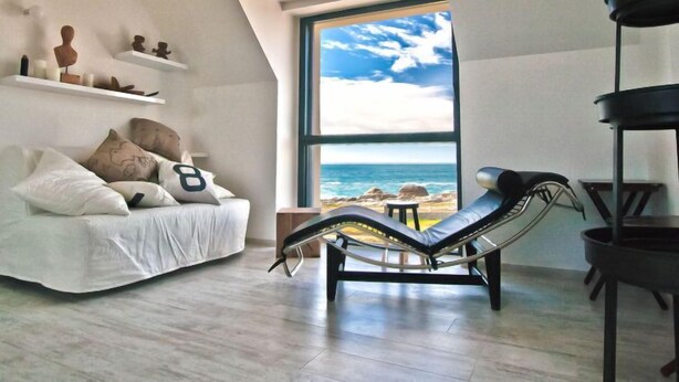 A modern living room with a white sofa, a lounge chair, and a large window overlooking the beach. - Le Ménez by Interhome (Penmarc'h)