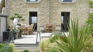 A stone building with two windows and a door, a patio with outdoor furniture, and a garden with various plants. - Le Ménez by Interhome (Penmarc'h)
