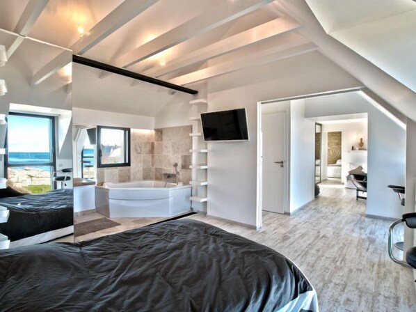 A modern bedroom with a large bed, a bathtub, a TV, and a view of the sea.