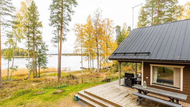 Una cabaña con terraza, una parrilla y vistas a un lago rodeado de árboles.