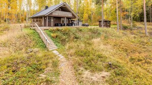 Una cabaña con porche y una estructura pequeña contigua, rodeada de un bosque de árboles de colores otoñales.