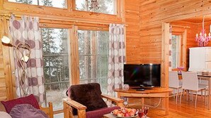 Interior acogedor de una cabaña de troncos, con un mueble de televisión de madera, un sillón mullido y un comedor con una mesa y sillas blancas.