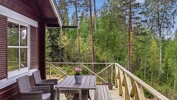 Terraza de madera con mobiliario de mimbre y una mesa, rodeada por una barandilla, con vistas a un área boscosa.