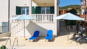 Et poolområde med to blå liggestole og to parasoller, en trappe til en balkon og udsigt til en bakke.