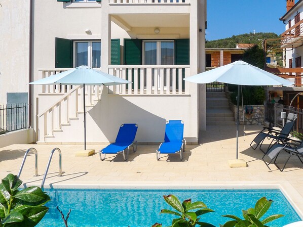 A poolside area with two blue lounge chairs and two umbrellas, a staircase leading to a balcony, and a view of a hillside. - Villa Zlatan by Interhome (Hvar/Stari Grad)