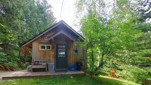 A small wooden cabin with a porch, a bench, and a trash bin.