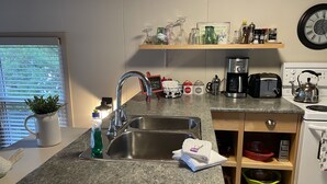 A kitchen with a granite countertop, stainless steel sink, and a coffee maker. There is a shelf above the sink holding various kitchen items and a clock on the wall.