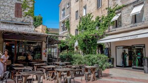 An outdoor café with wooden tables and chairs, a stone building with ivy, and a clothing store named 'Carioca Plage'. - Résidence Cita di Sali by Interhome (Porto Vecchio)