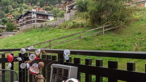 Un balcó amb vista a un poble situat en un turó, una barana de fusta rústica i una taula amb una tetera, una tassa i objectes decoratius.
