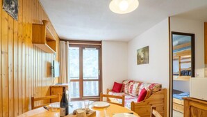 A cozy dining area with a wooden table set for two, a sofa with a colorful cushion, and a bunk bed in the background. - Astragale 412 A by Interhome (Les Menuires)