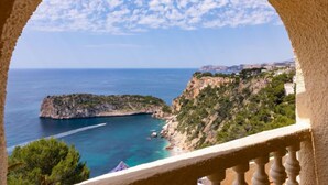 A coastal view through an arched window overlooking a clear blue sea, rocky cliffs, and a distant town. - Vacation home Igor by Interhome (Jávea)