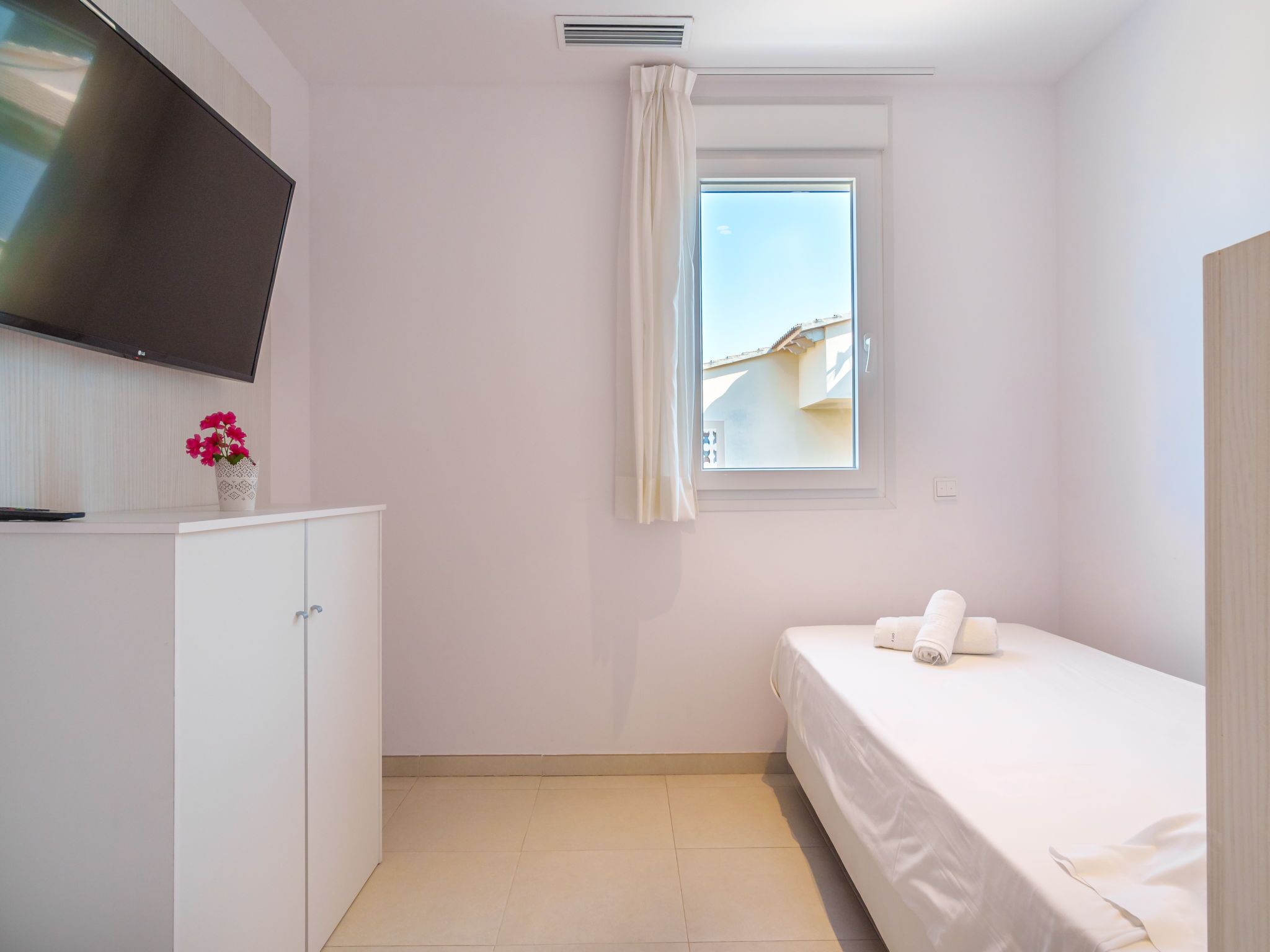 A small, clean room with a bed, a TV mounted on the wall, a window with curtains, and a cabinet with a vase of flowers.