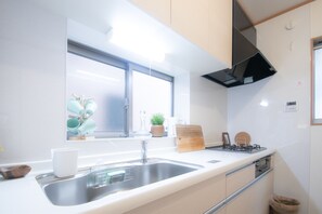 A modern kitchen with a stainless steel sink, a window with a view, a stove, and a countertop with various items. - AMP FLAT Haruyoshi A (Fukuoka)