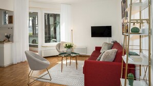 A modern living room with a red sofa, a grey armchair, and a glass coffee table. There is a TV mounted on the wall, a bookshelf, and a window with a view of palm trees.