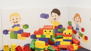 Una habitació amb una paret blanca i un terra amb moqueta, amb un gran munt de blocs de construcció de colors i tres personatges de dibuix animat.
