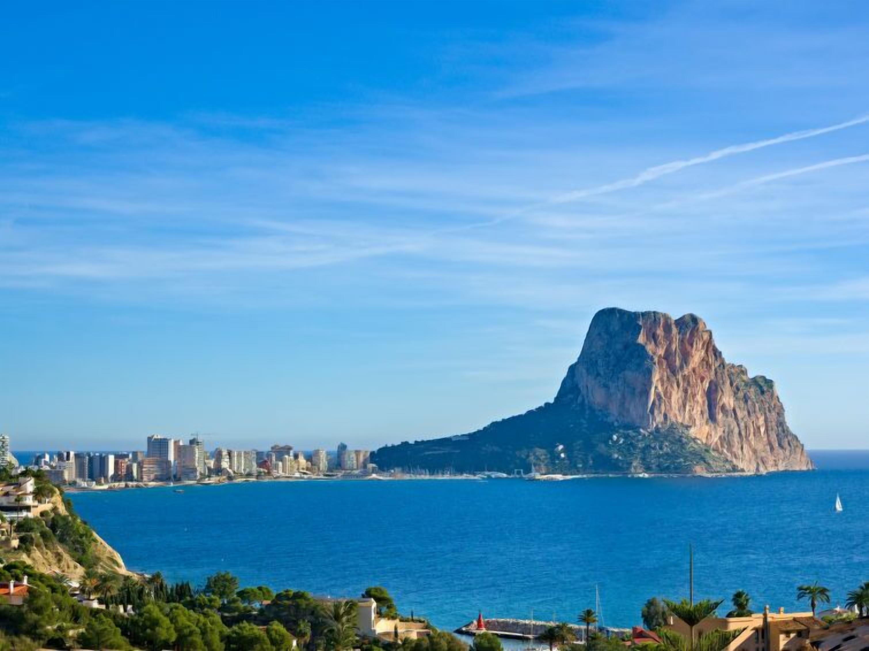 A coastal cityscape with a prominent rocky hill, a clear blue sky, and a calm sea.
