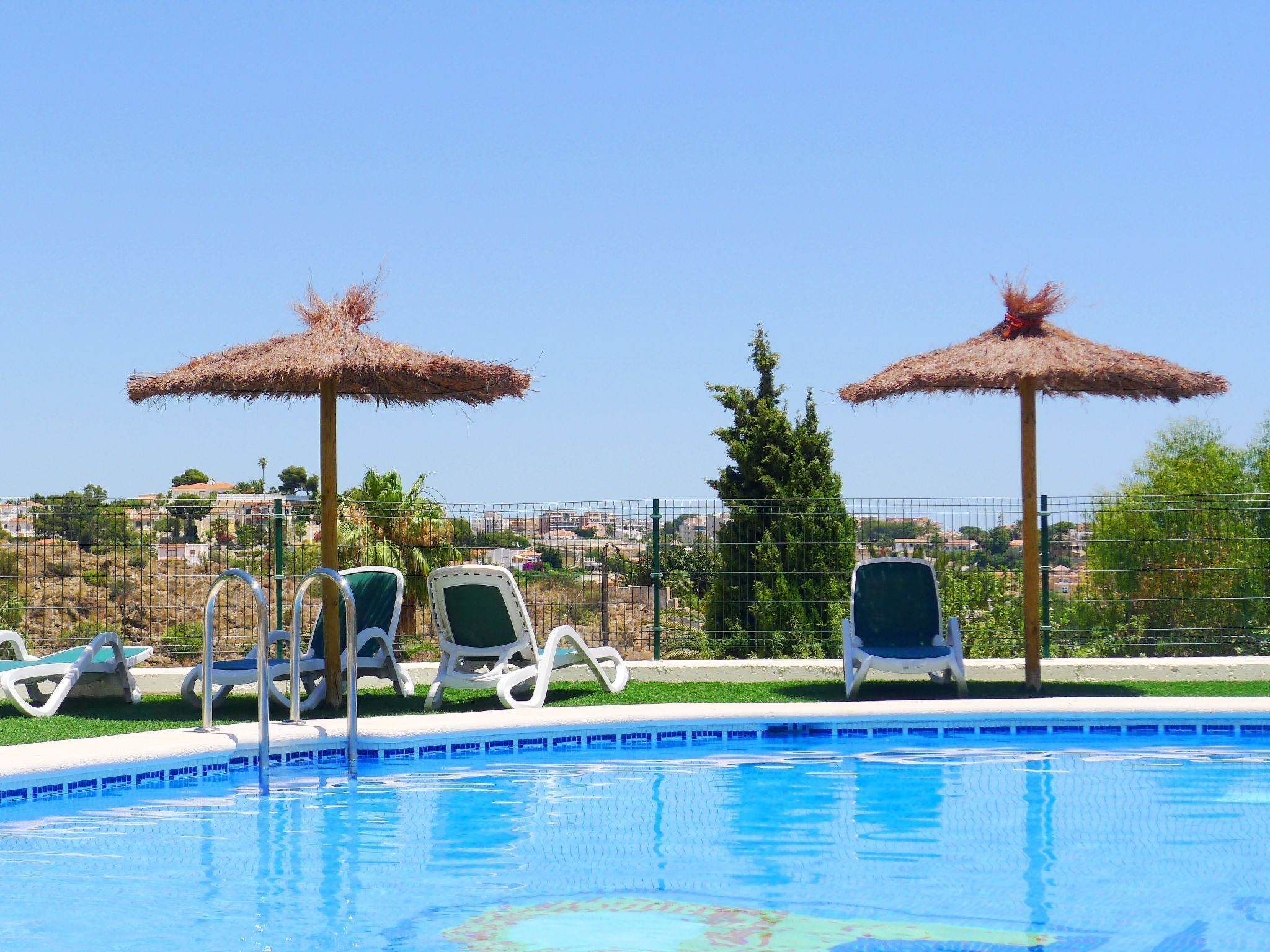 Ein Swimmingpool mit Liegestühlen und Strohdächern, umgeben von einem Zaun und Grünpflanzen.