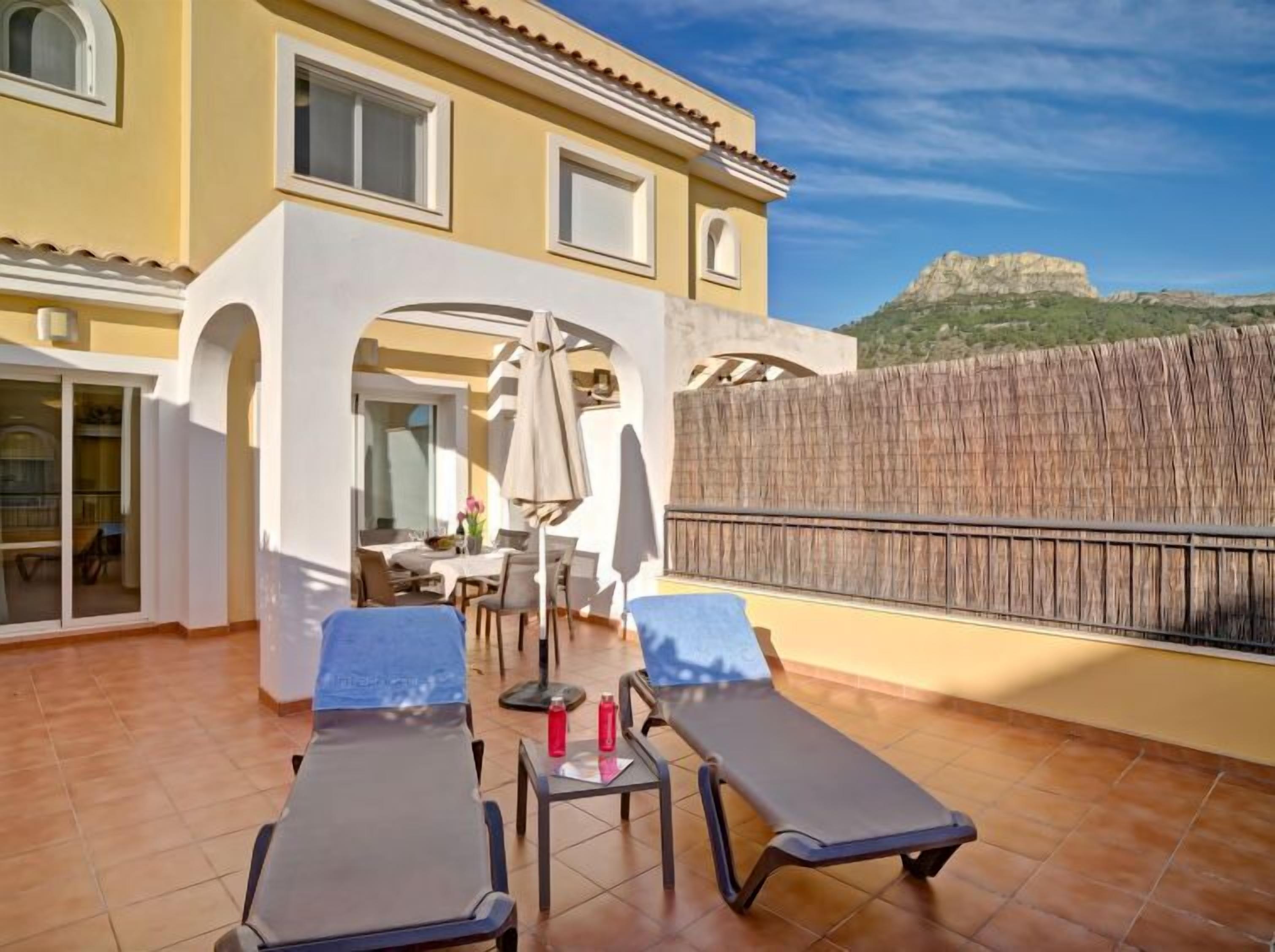 Ein gelbes Gebäude mit Terrasse, zwei Liegestühlen, einem Tisch und Blick auf einen Berg.