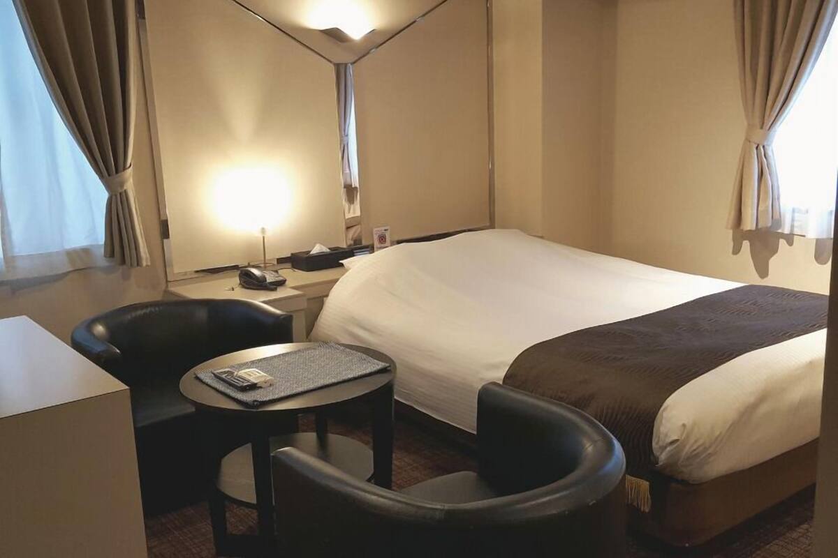 A hotel room with a bed, a desk, a chair, a small table, and a lamp.