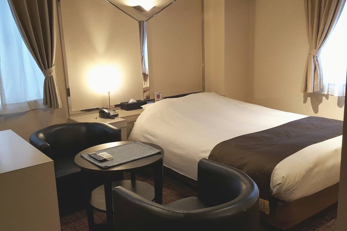 A hotel room with a bed, a desk with a telephone, a chair, a small round table, and a lamp.