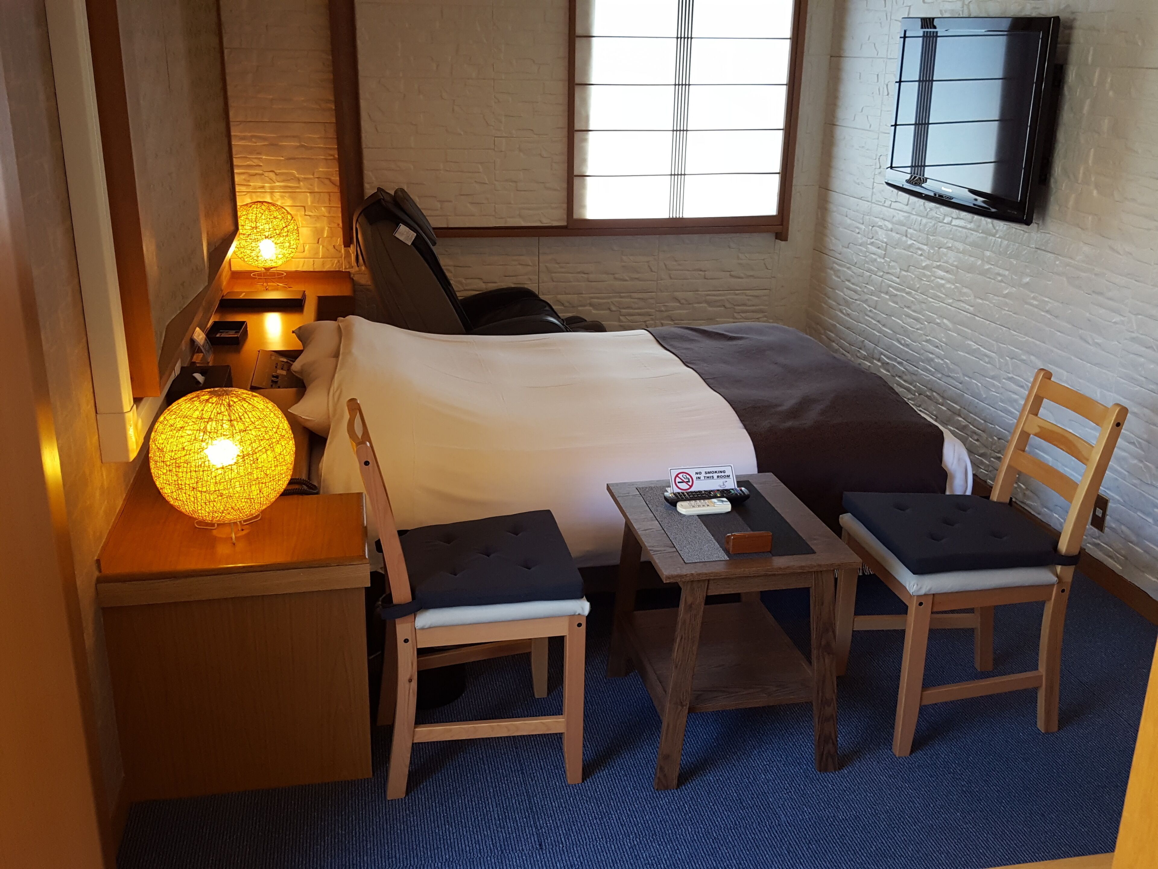 Habitació d'hotel amb llit, taulella de nit, làmpada, cadira i una taula petita amb un senyal.