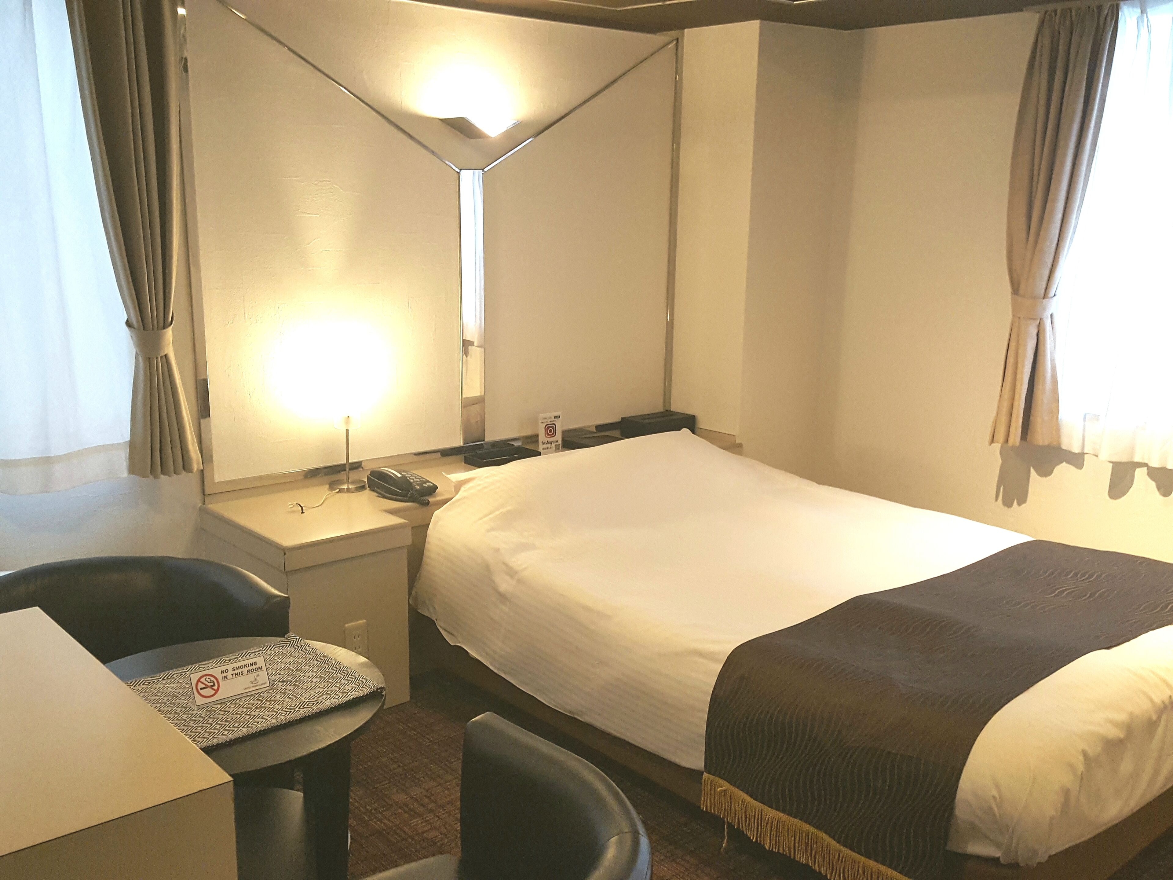 Habitació d'hotel amb un llit, una taula de treball amb un telèfon, una cadira i una taula petita amb un senyal de 'No Fumar'.