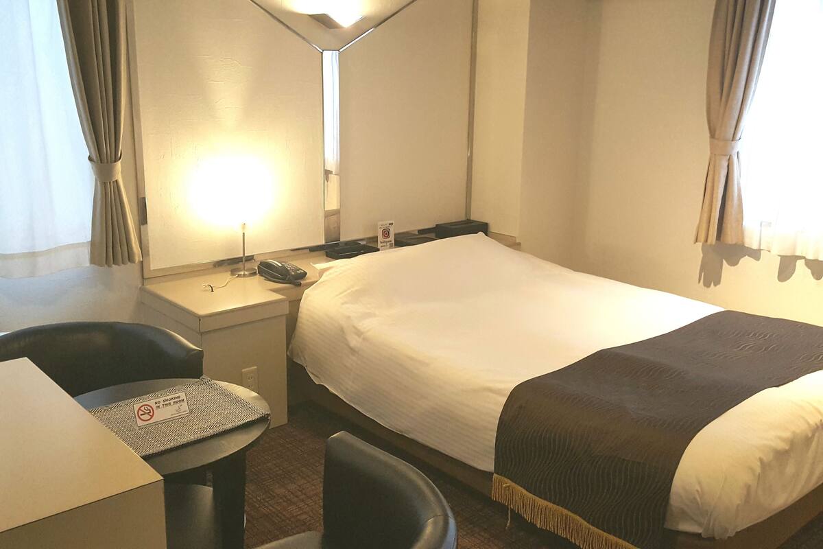 A hotel room with a bed, a desk with a telephone, a chair, and a small table with a 'No Smoking' sign.
