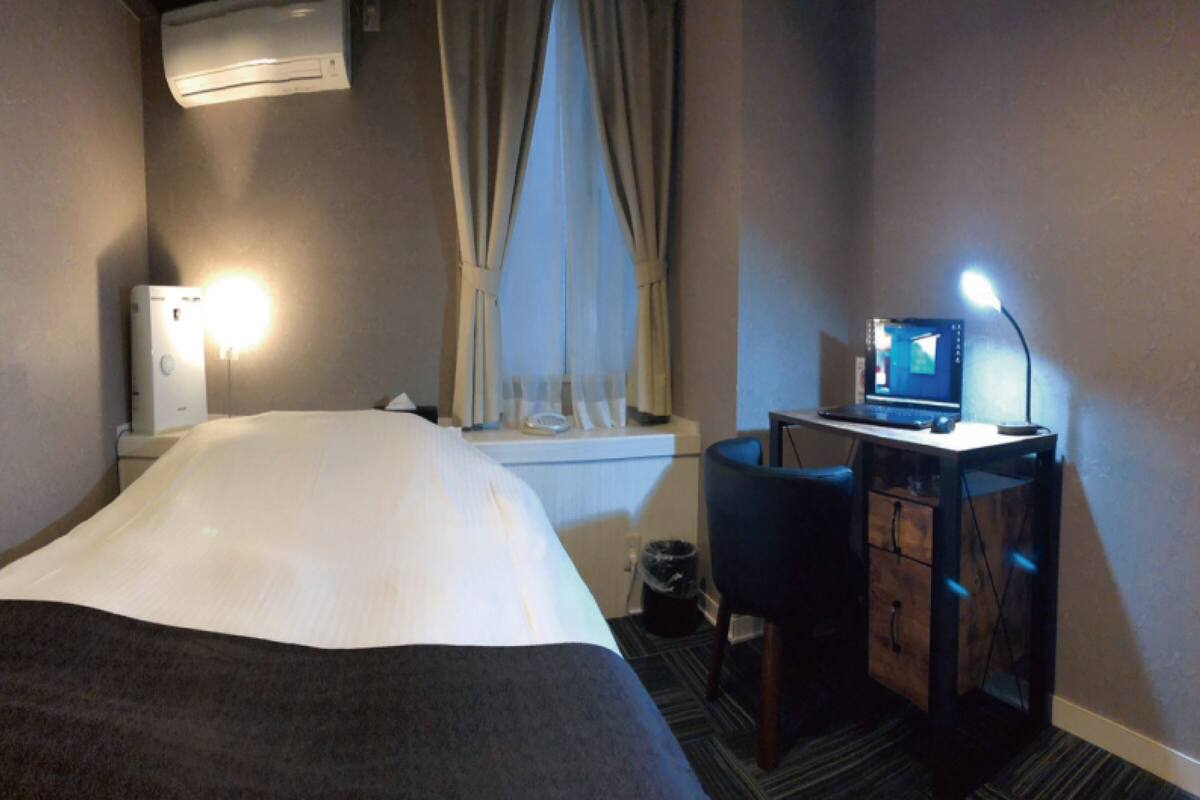 A hotel room with a bed, a desk with a computer, a chair, a window with curtains, and an air conditioning unit.