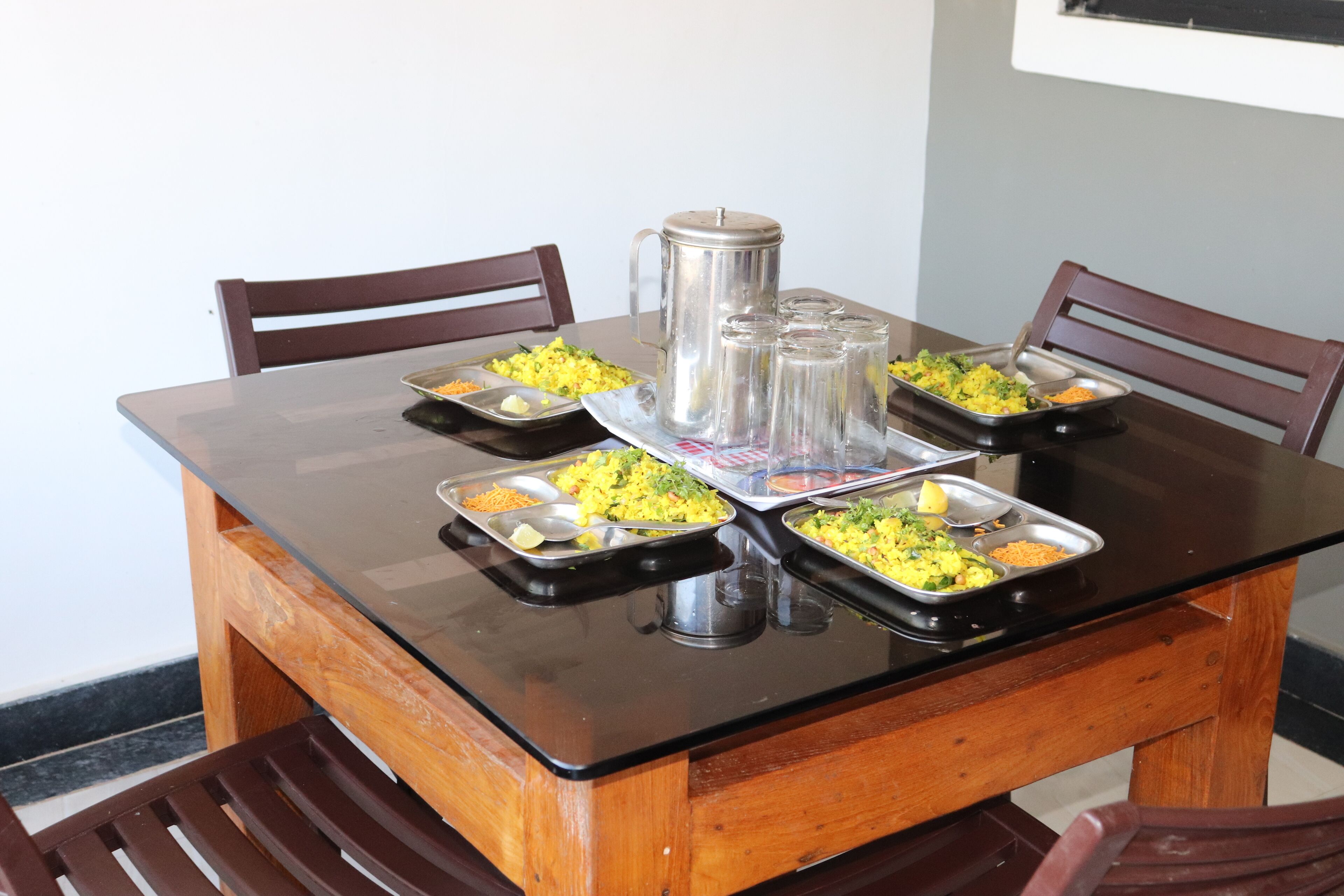 Une table à manger avec quatre plateaux en métal contenant des plats, une cafetière en acier inoxydable et quatre bouteilles d’eau en verre.