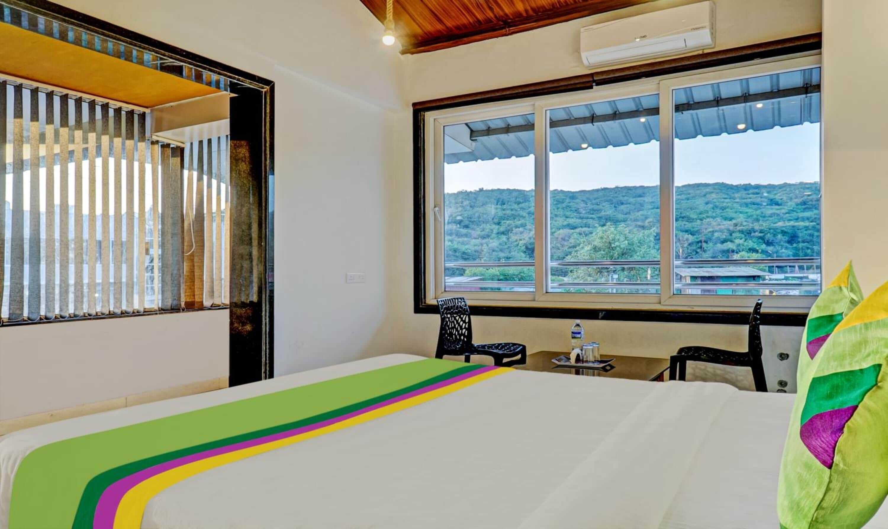 A bedroom with a large window, a bed with a colorful bedspread, a chair, and a small table with a water bottle.