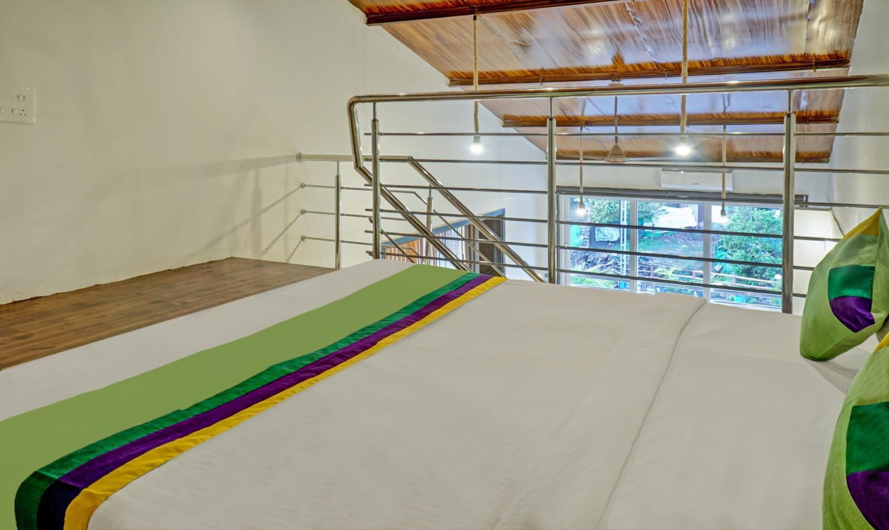 A bedroom with a bed, a wooden floor, a metal railing, and a view of greenery outside.