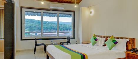 Quarto de hotel com uma cama grande, cabeceira de madeira, escrivaninha com cadeira, janela com vista para áreas verdes e ar condicionado.