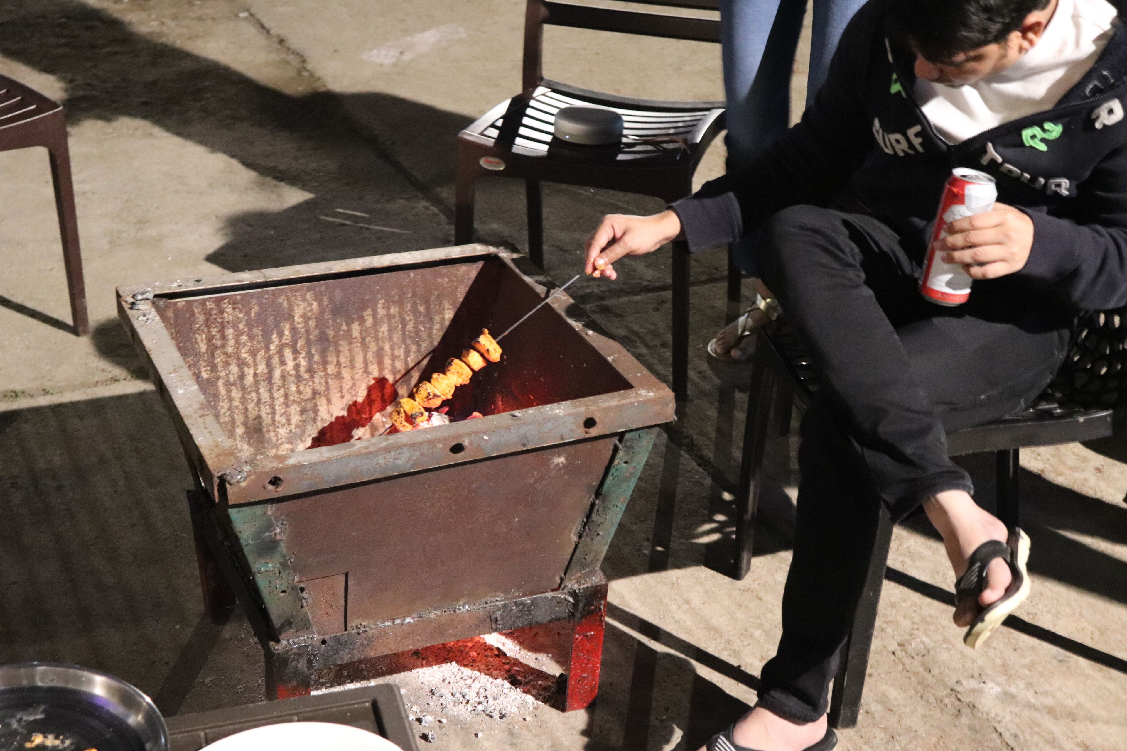 Eine Person grillt im Freien, sitzt auf einem Stuhl und hält eine Dose Getränk in der Hand.