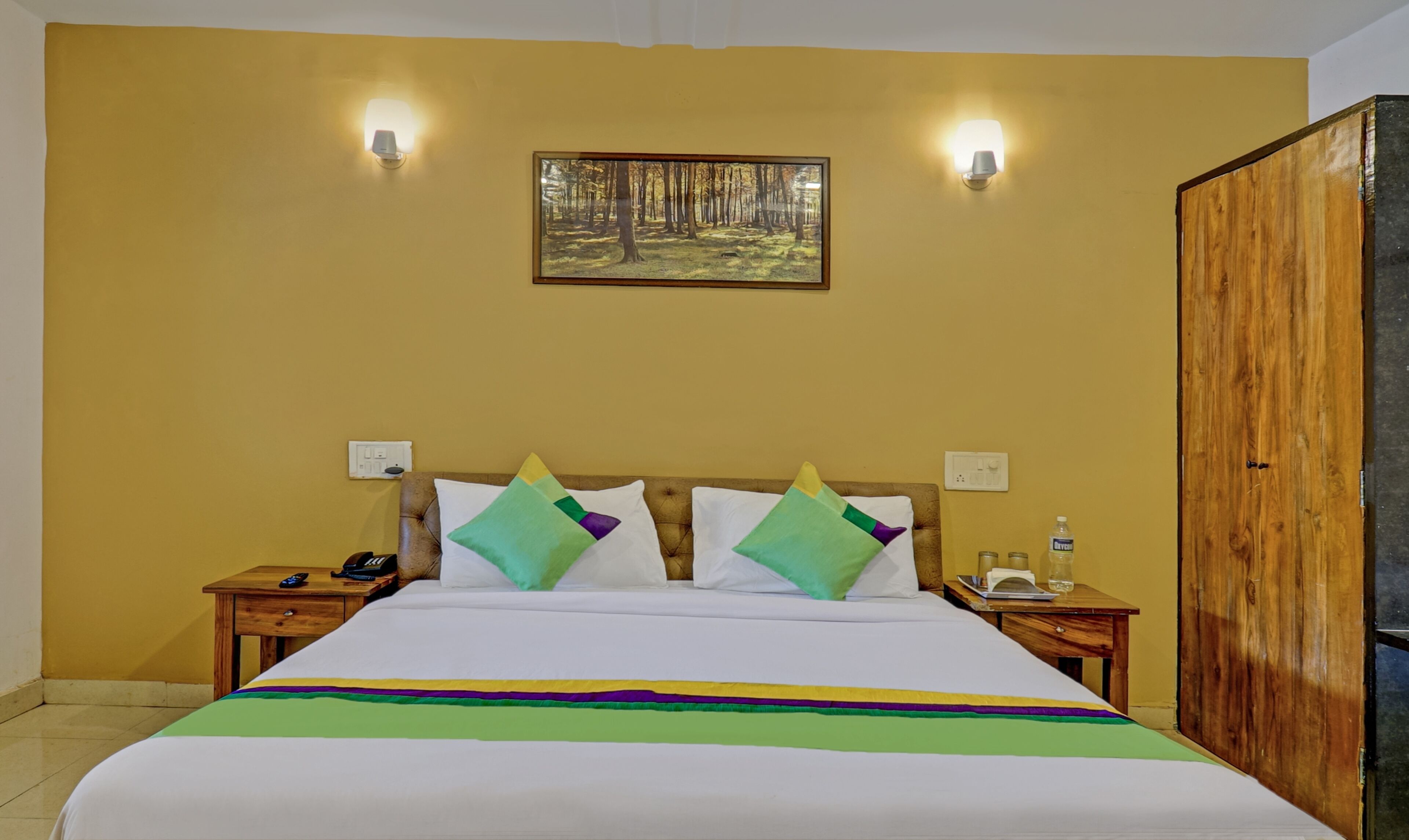 A hotel room with a bed, two nightstands, a wooden wardrobe, and a forest-themed painting on the wall.