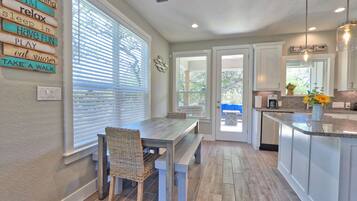 A modern kitchen with a dining area, featuring a wooden table and chairs, a built-in island with a sink, and a view through the window.