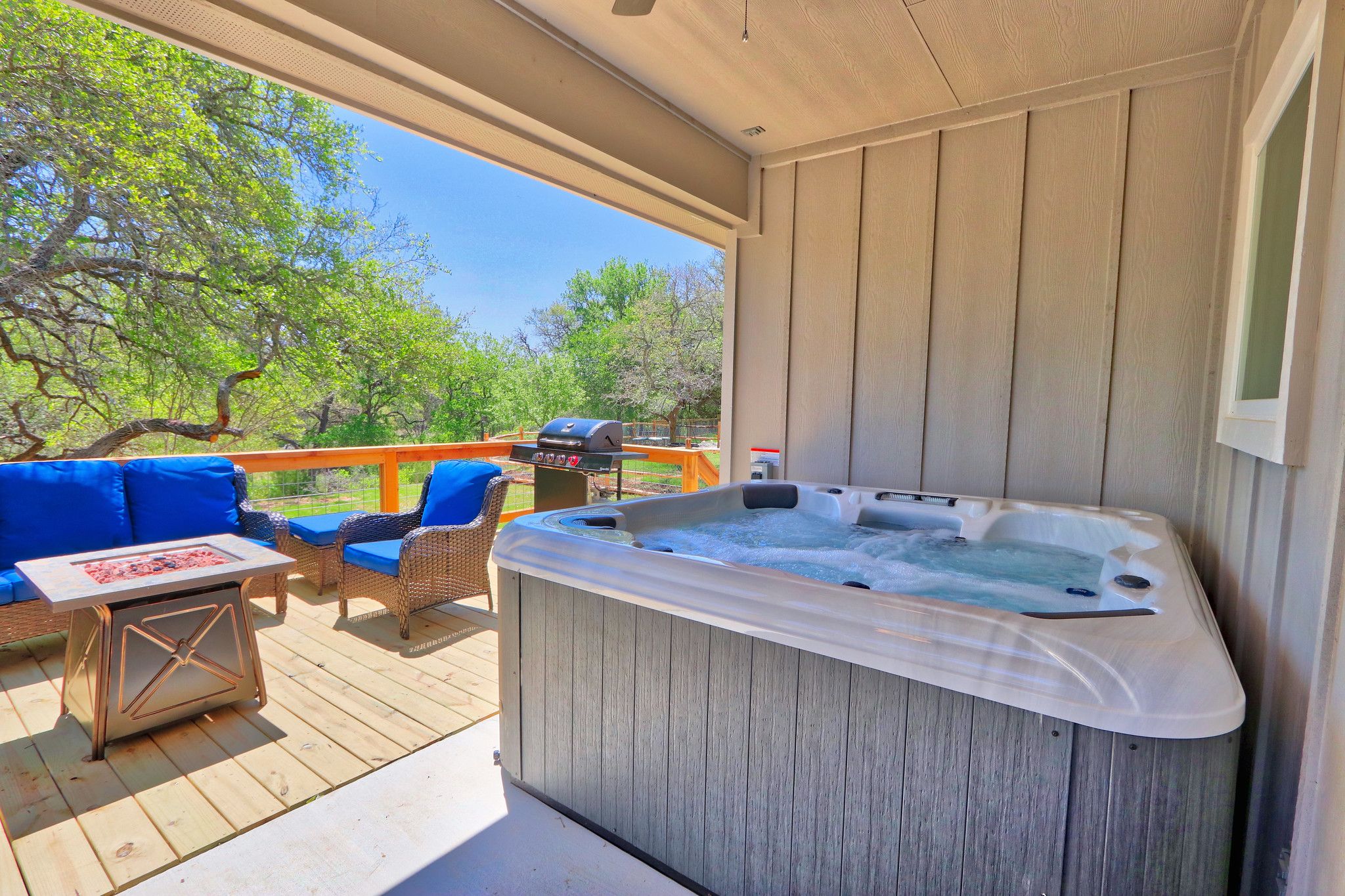 A hot tub on a wooden deck with blue outdoor furniture and a view of trees.