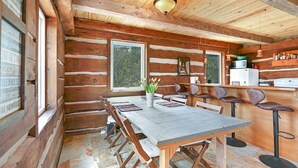 Interieur van een houten hutje met een eettafel, stoelen, een keuken en een raam met uitzicht op bomen.