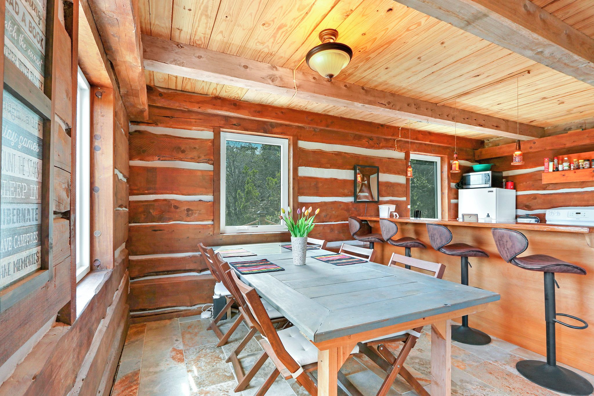 Interior rústico de una cabaña con una mesa de comedor de madera, sillas, una zona de cocina con un refrigerador y un ventanal con vistas a árboles.