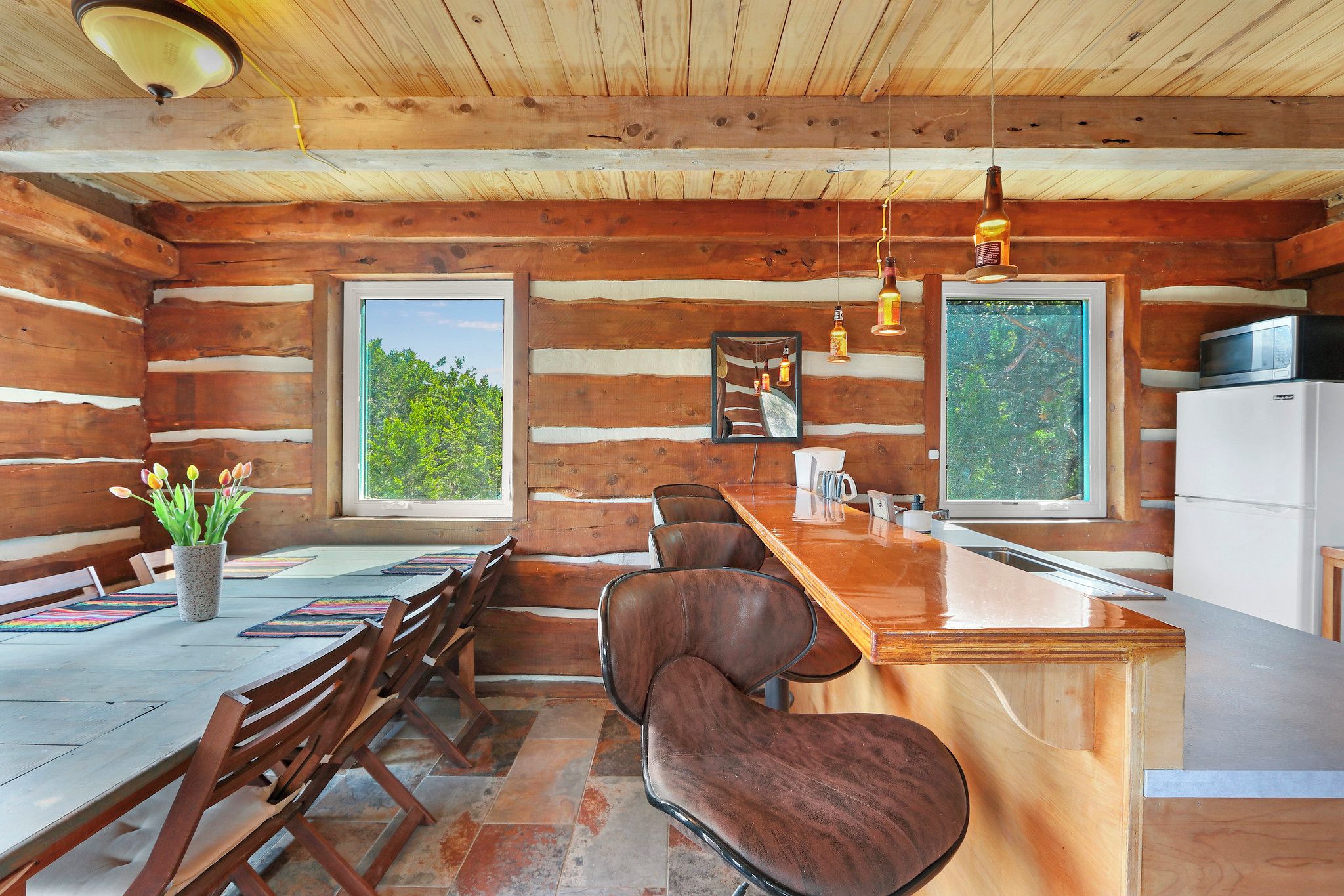 A wooden cabin interior with a dining area, kitchen, and large windows.