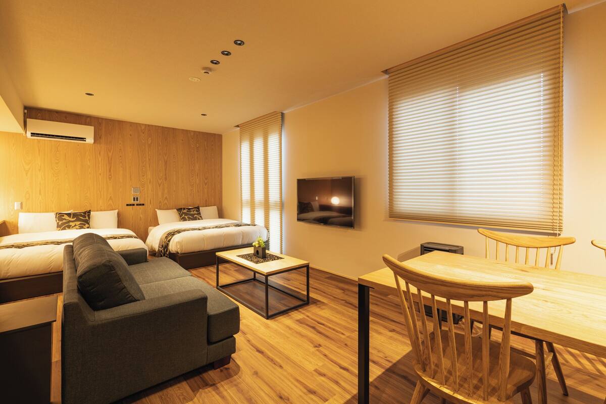 A modern hotel room with two beds, a sofa, a dining table, and a TV.