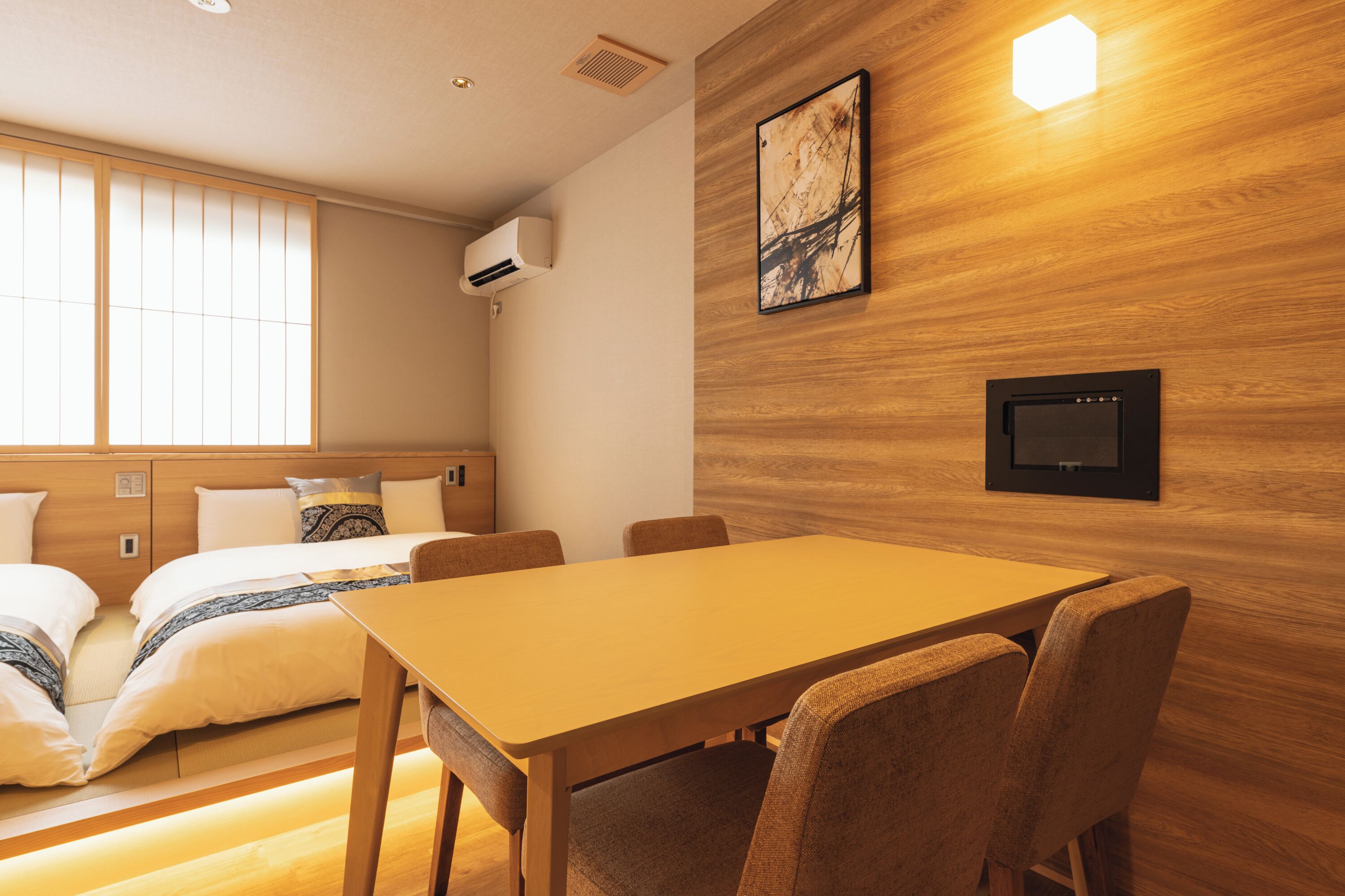 Quarto de hotel com parede de madeira, cama com roupa de cama branca, mesa com cadeiras e televisão montada na parede.