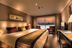 Habitación de hotel con una cama grande, un escritorio con televisor, una silla y vistas a la ciudad al atardecer.