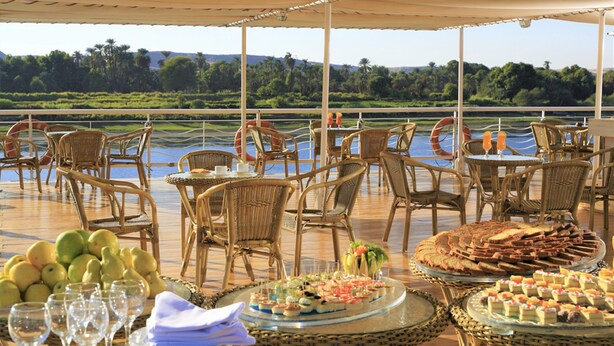 Un área de comedor al aire libre con sillas y mesas de mimbre, un bufé con diversos postres, y vistas a un río y follaje en el fondo.