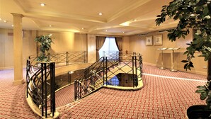 Un vestíbulo de hotel con alfombra roja estampada, una escalera central con barandilla negra y una planta grande en primer plano.