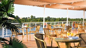 An outdoor deck with wicker furniture and tables, overlooking a body of water with a view of greenery in the distance. - JAZ Crown Jubilee Nile Cruise - Every Thursday from Luxor for 07 & 04 Nights - Every Monday from Aswan for 03 Nights (Luxor)