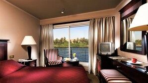 A hotel room with a large bed, a desk with a television, a chair, and a view of a body of water and greenery. - JAZ Crown Jubilee Nile Cruise - Every Thursday from Luxor for 07 & 04 Nights - Every Monday from Aswan for 03 Nights (Luxor)