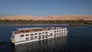 Un gran barco de paseo fluvial con varios niveles y cubierta superior protegida, navegando por un río con un paisaje desértico al fondo.