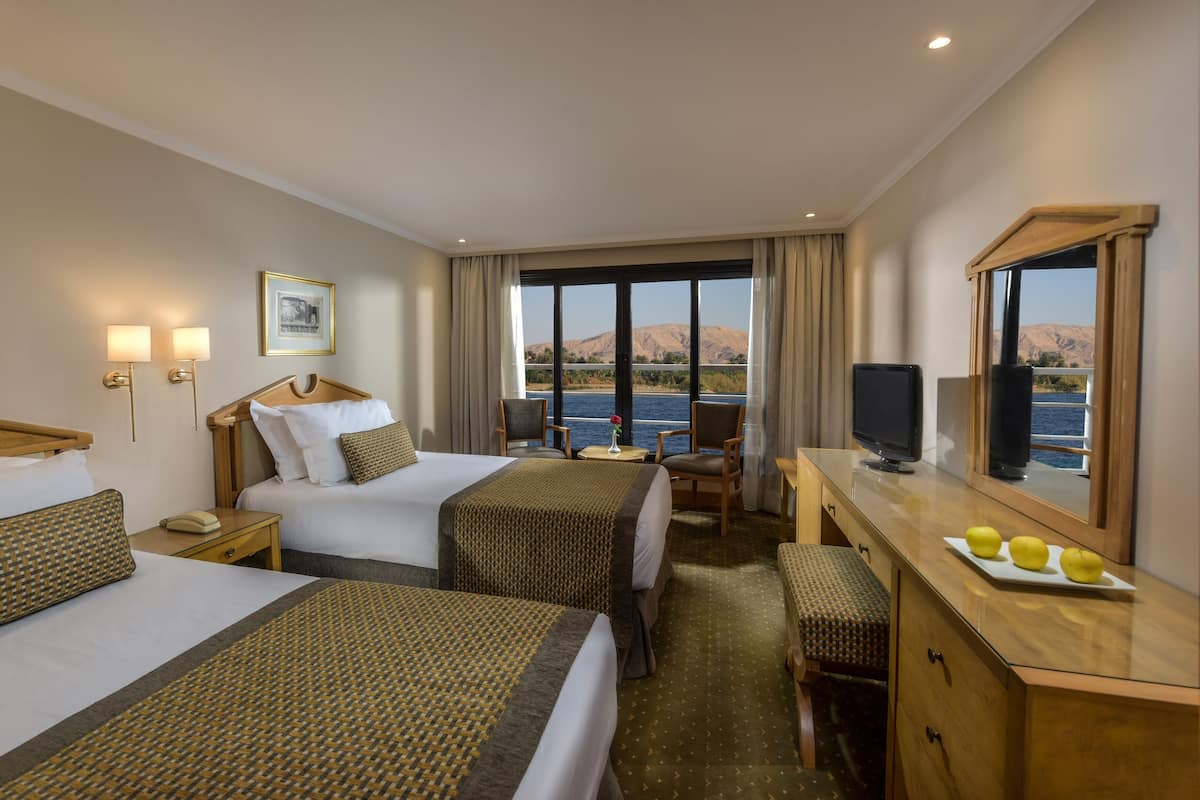 A hotel room with two beds, a desk with a television, a small table with a vase, and a view of a body of water and mountains.